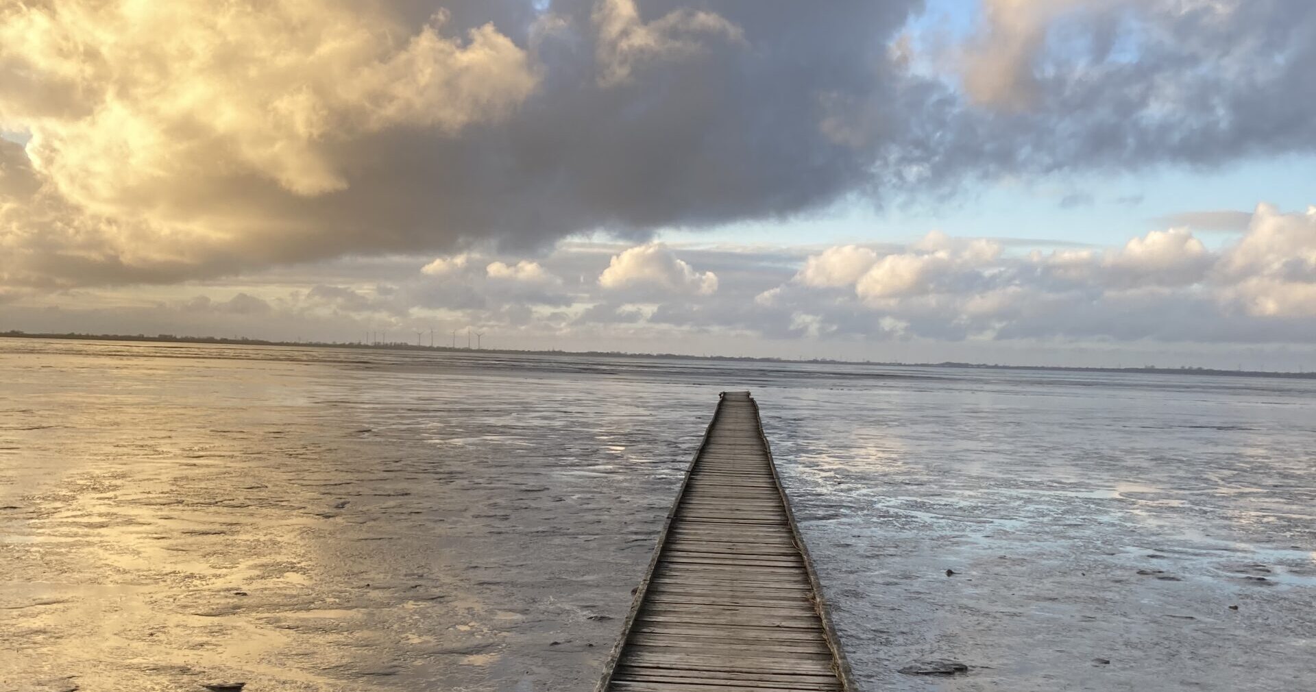 Holzsteg führt über eine weite Wattfläche unter einem bewegten Wolkenhimmel